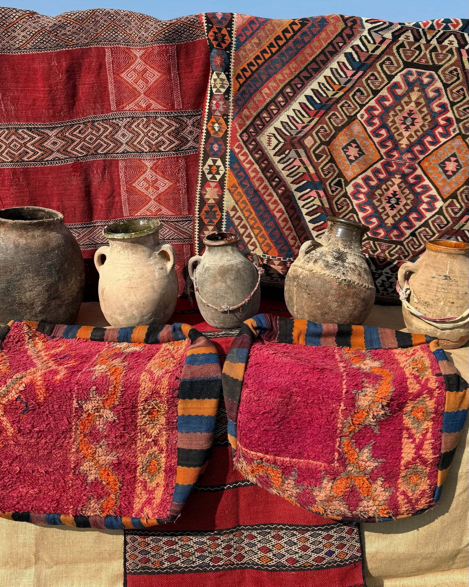 Antique terracotta pots and handwoven Moroccan textiles with vibrant geometric patterns displayed outdoors.